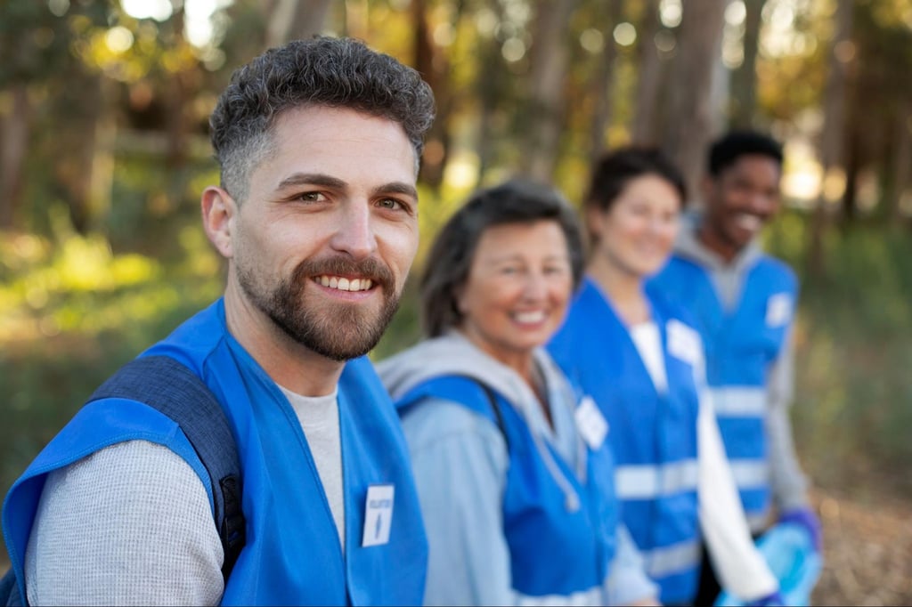 A man in a blue volunteer vest smiles at the camera with his team standing in a line behind him, slightly out of focus, in a sunny park setting.