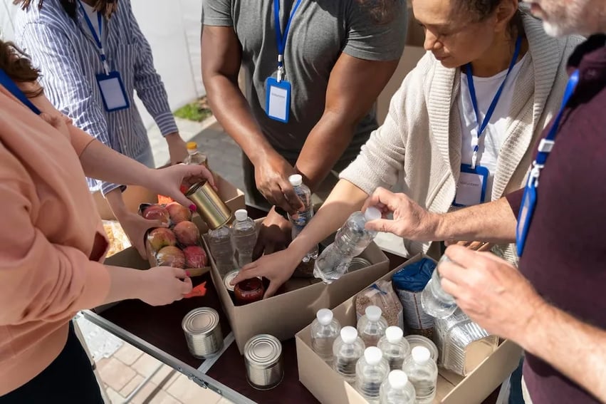 A group of volunteers wearing ID badges packing cardboard boxes with bottled water, canned goods, and fresh fruit at an outdoor relief event.