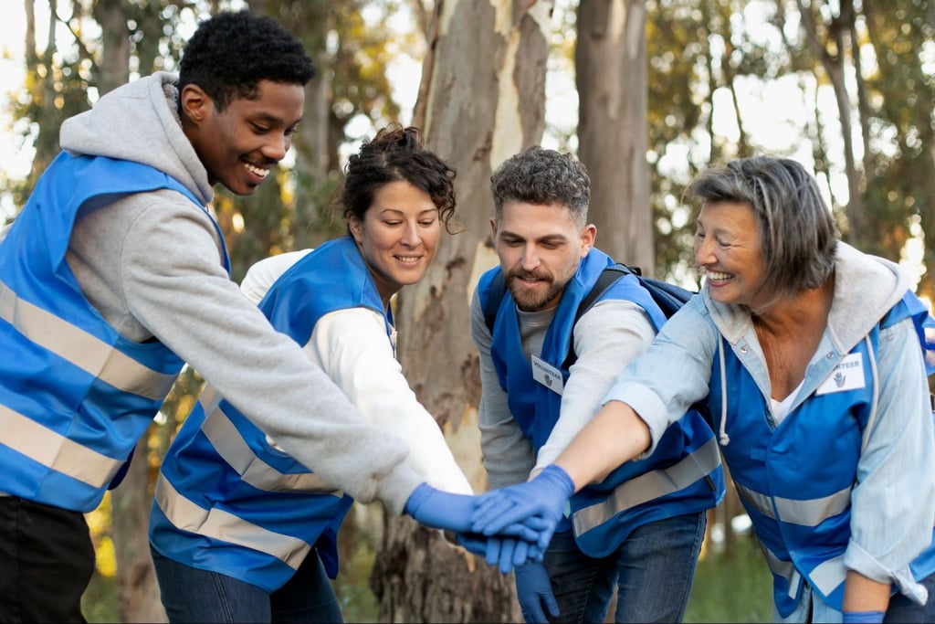 Smiling team of adult volunteers in blue vests stacking hands in a forest setting, showing teamwork and community spirit.