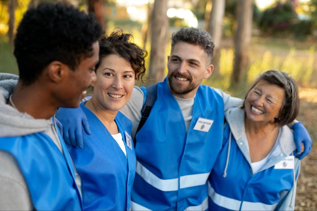 A diverse group of volunteers wearing blue vests stand arm-in-arm, smiling and talking in an outdoor wooded area.